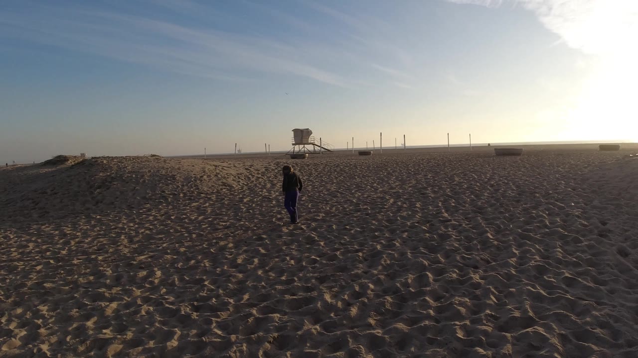 At Bolsa Chica, California, the adult and child are positioned right of center, with open sand, a lifeguard tower, and a bright low sun haze across the background.