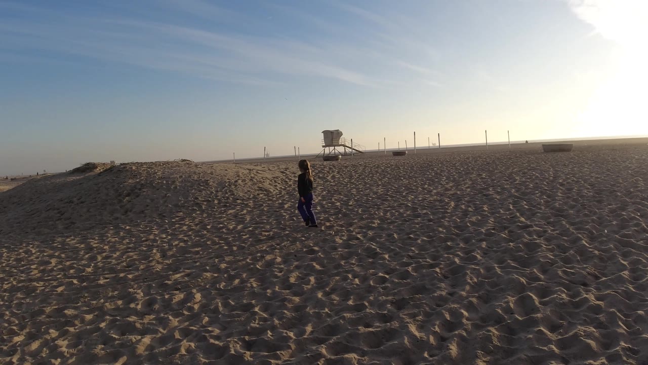 Adult and child walking across sand with lifeguard tower in the distance.