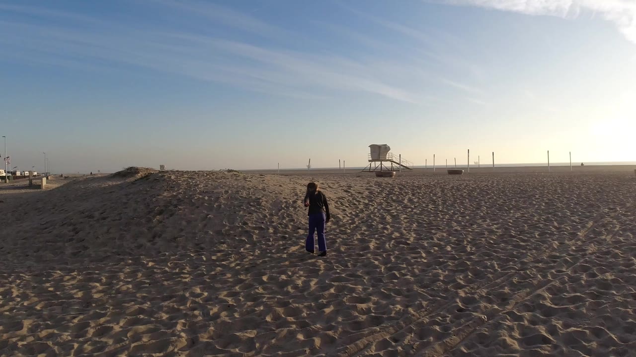 At Bolsa Chica, California, a small girl stands on rippled sand with a lifeguard tower and bright low sun near the horizon.