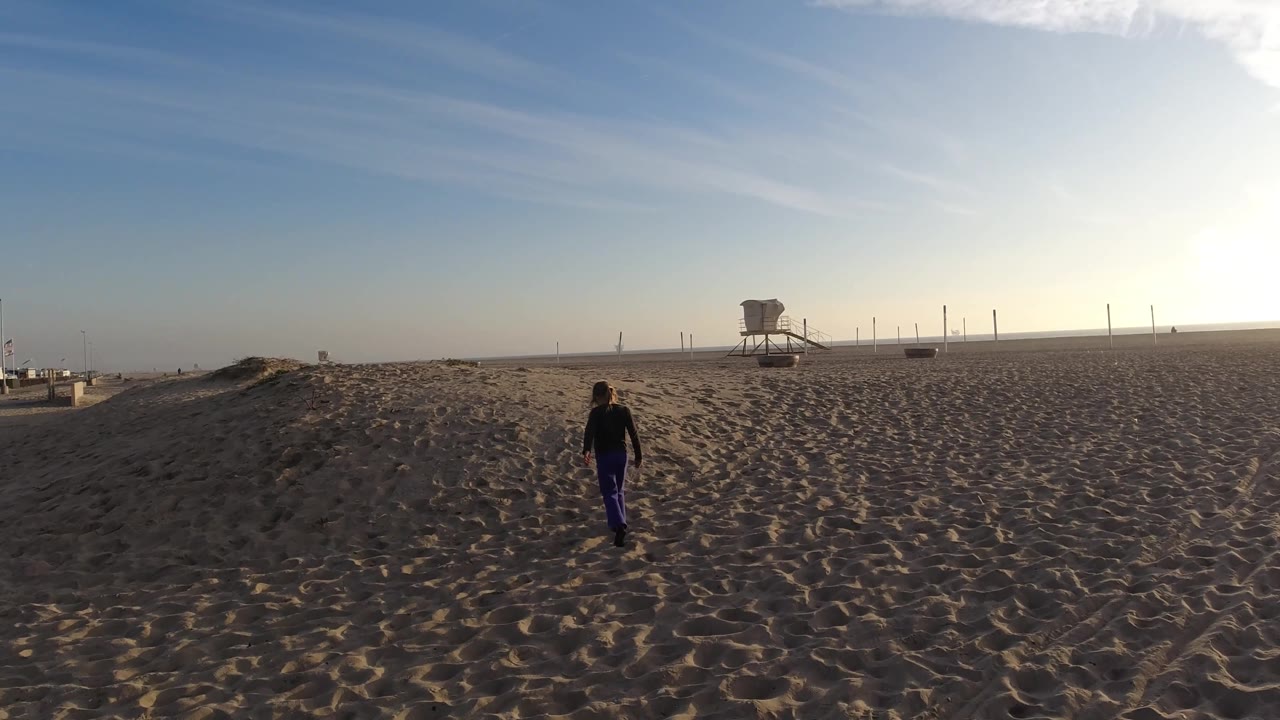 At Bolsa Chica, California, the girl appears farther right on the sand while the lifeguard tower and fence line remain visible against the bright sky.