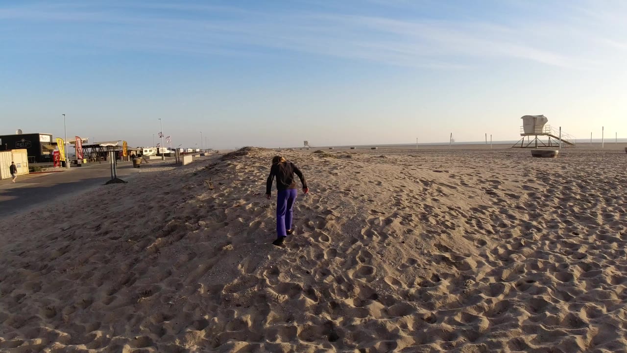 At Bolsa Chica, California, a young girl in purple pants walks away across a sandy dune area with beach kiosks at left and a lifeguard tower in the distance.
