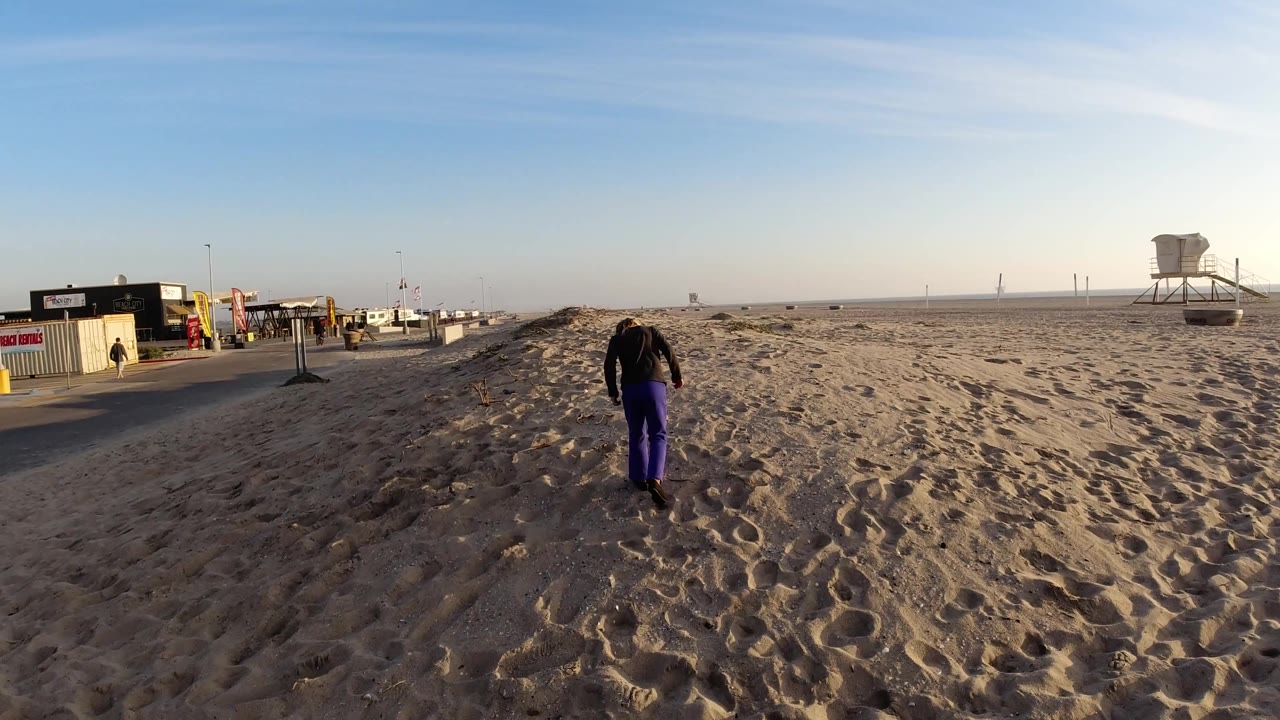 A young girl walks farther across the Bolsa Chica sand, with small beach buildings along the left side and a raised lifeguard tower near the right horizon.