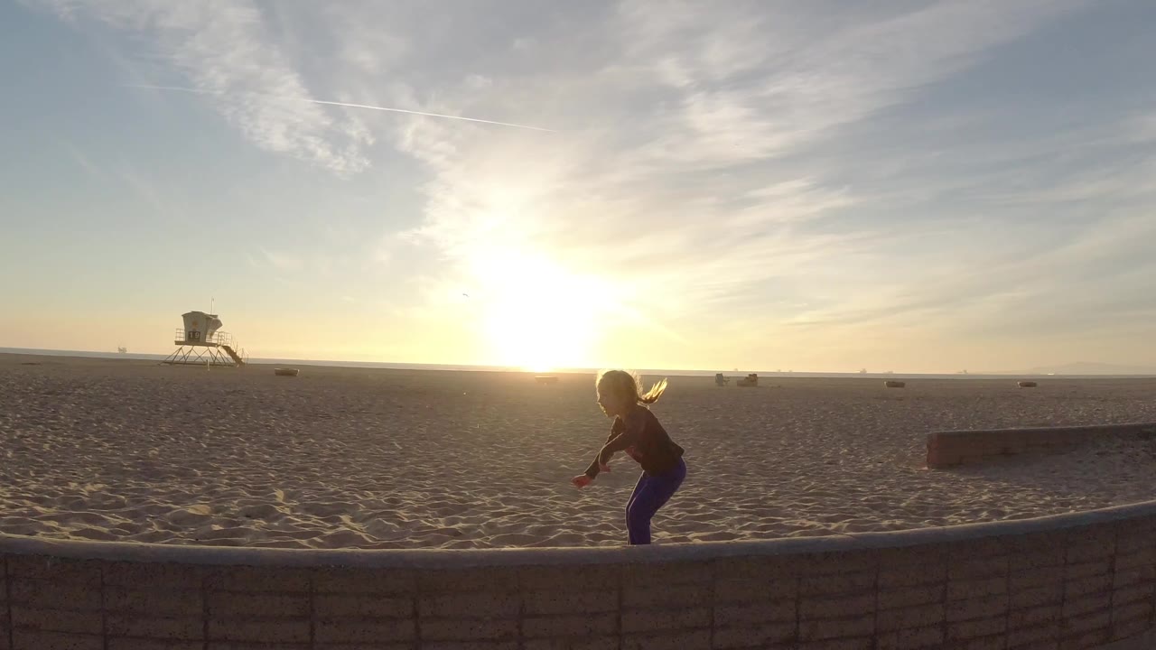 At Bolsa Chica, California, a young ponytailed girl balances barefoot on a low wall while the sun sits low over the sand and a lifeguard tower stands in the distance.