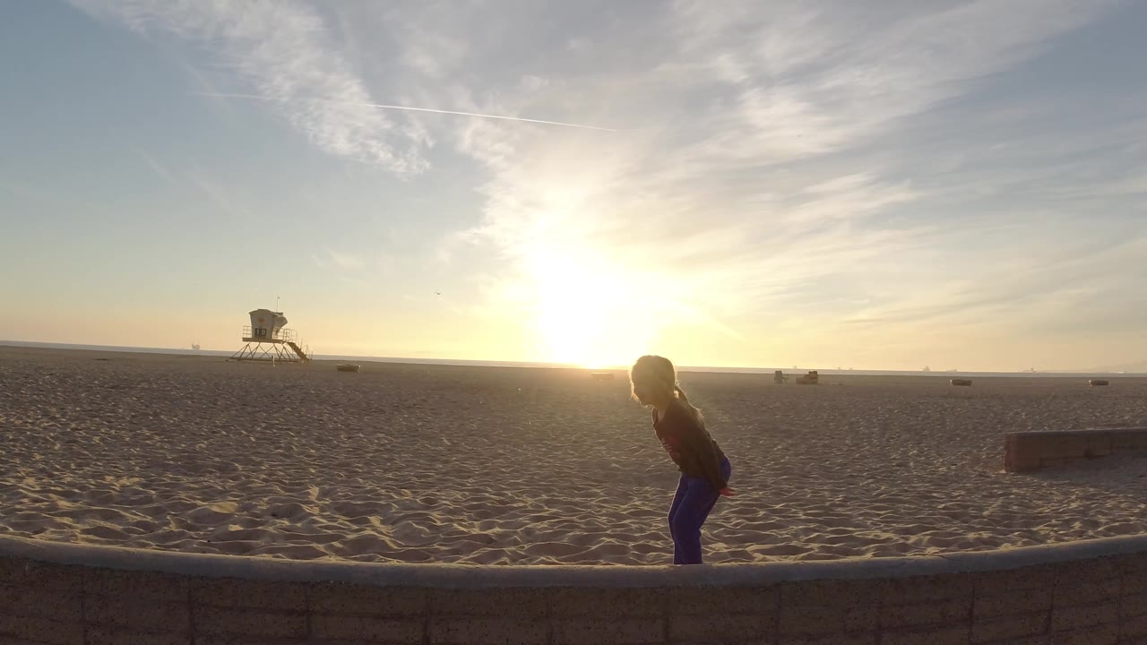 At Bolsa Chica, California, the girl moves farther right on the low wall, silhouetted by the low sun with a lifeguard tower and sparse beach activity behind her.