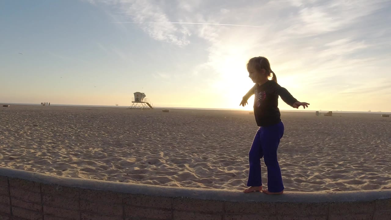 The child continues along the low wall in Bolsa Chica, California, arms extended while the sun glows behind the shoulder and sand fills the background.