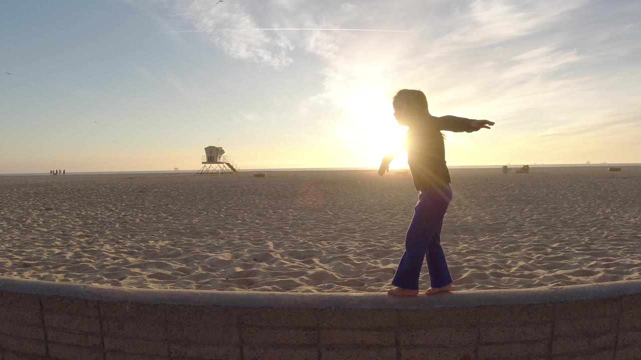 A backlit child balances on the low wall in Bolsa Chica, California, with the bright sun partly hidden by the body and the lifeguard tower set far across the sand.