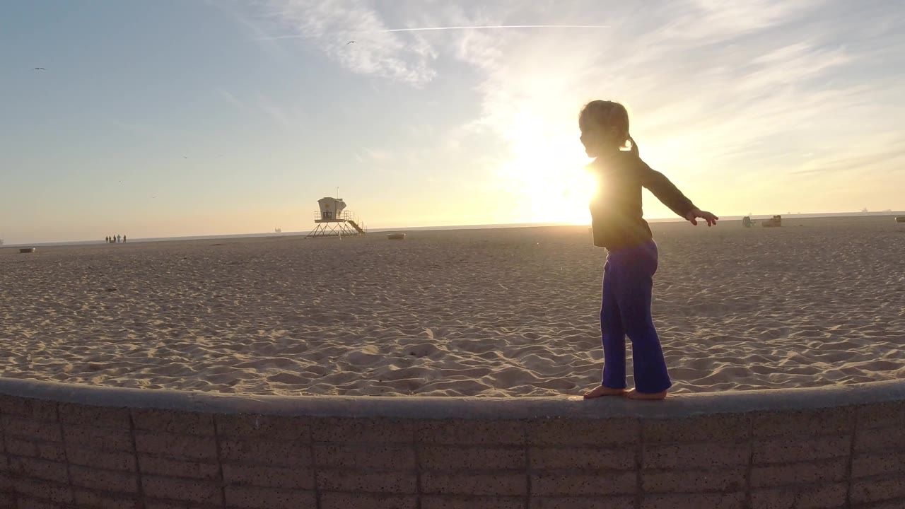 Child in profile balancing along a beach wall in front of open sand.