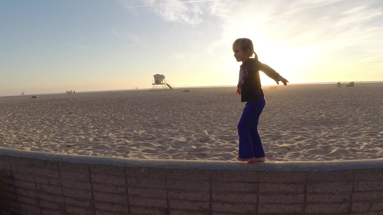A young girl balances on a thin low wall at Bolsa Chica, with open sand, a lifeguard tower, distant beachgoers, and sunset backlight behind her.