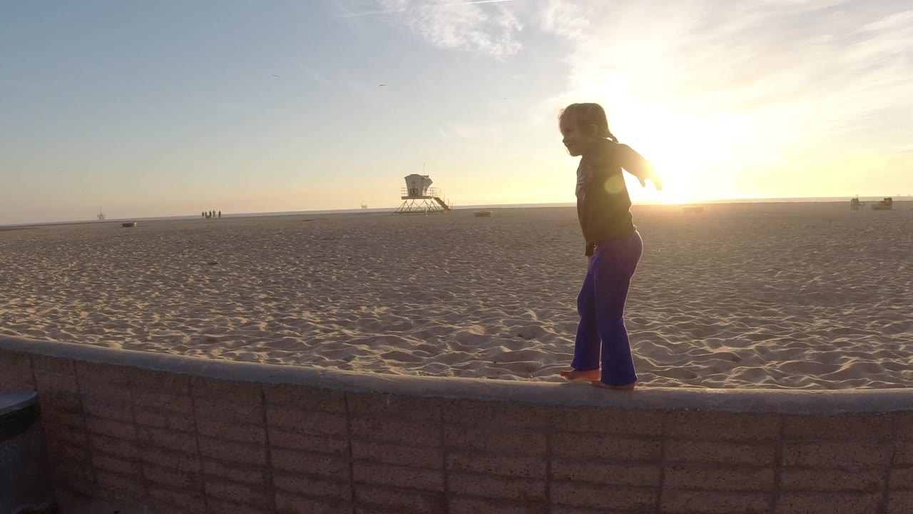 The girl nears the right side of the wall at Bolsa Chica, partly silhouetted by the low sun with the ocean horizon and small distant figures behind her.
