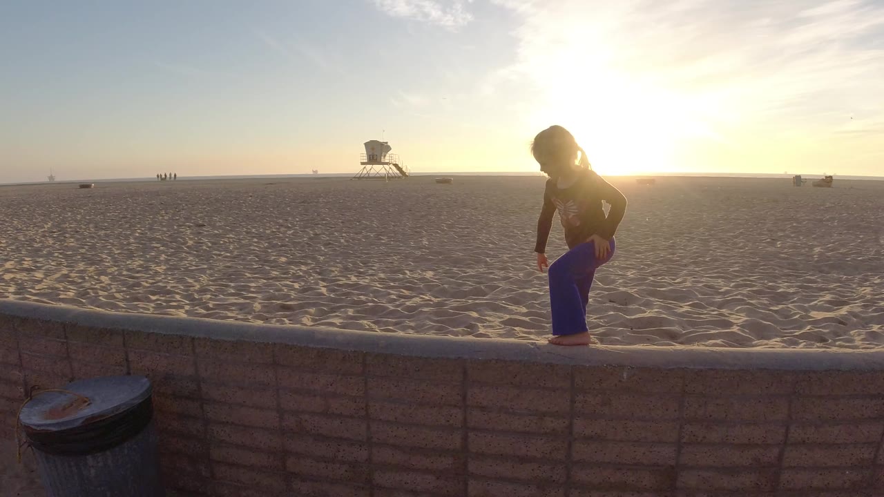 A young girl stands on a low wall at Bolsa Chica Beach with the sun behind her, textured sand beyond, a distant lifeguard tower, and small figures near the horizon.