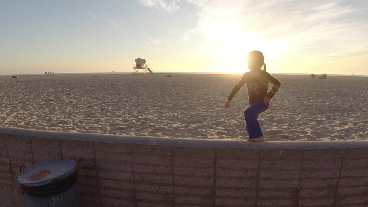 A young girl steps along the low wall at Bolsa Chica Beach, her head and shoulders silhouetted against the bright sun while the open sand and lifeguard tower stay in the background.