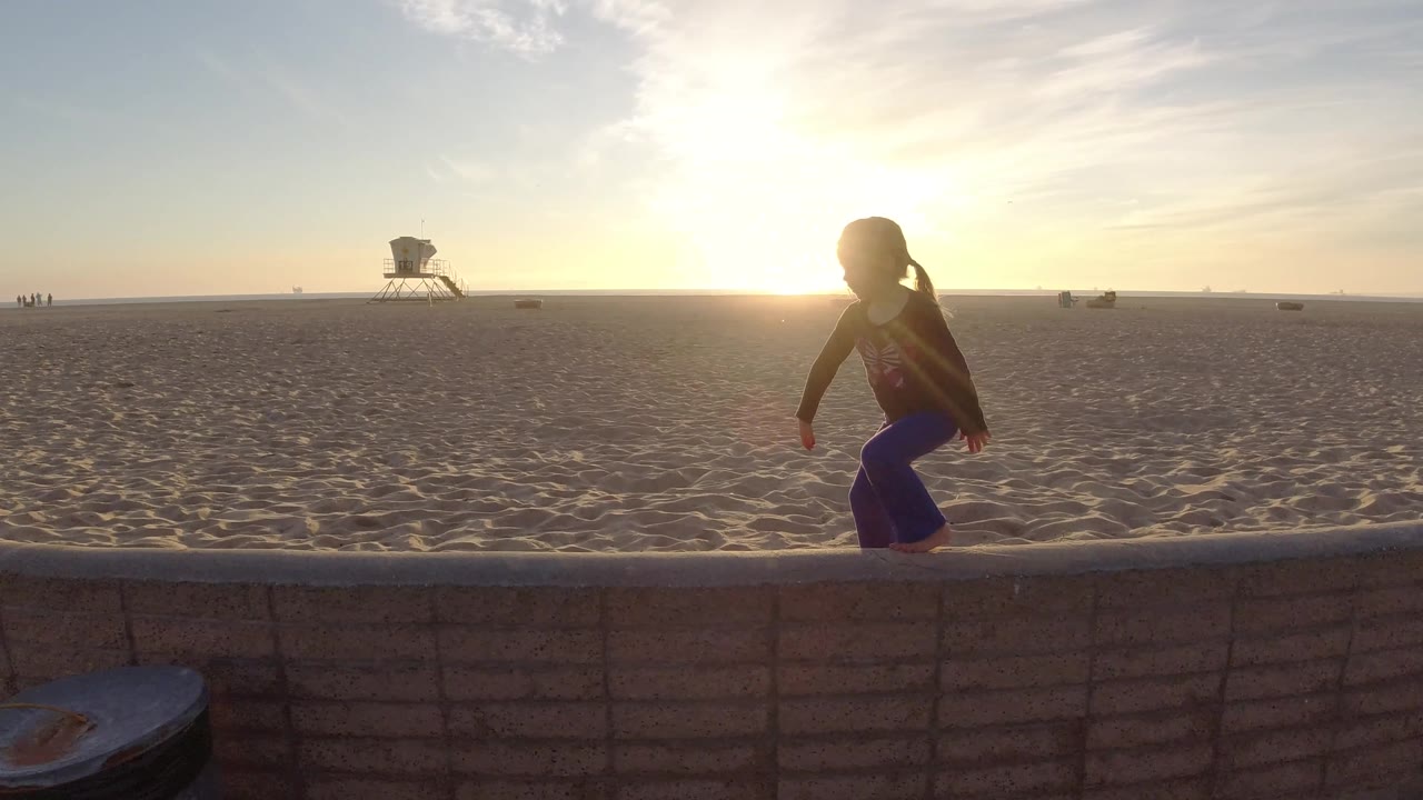 Child moving left on a wall beside a wide sandy beach.