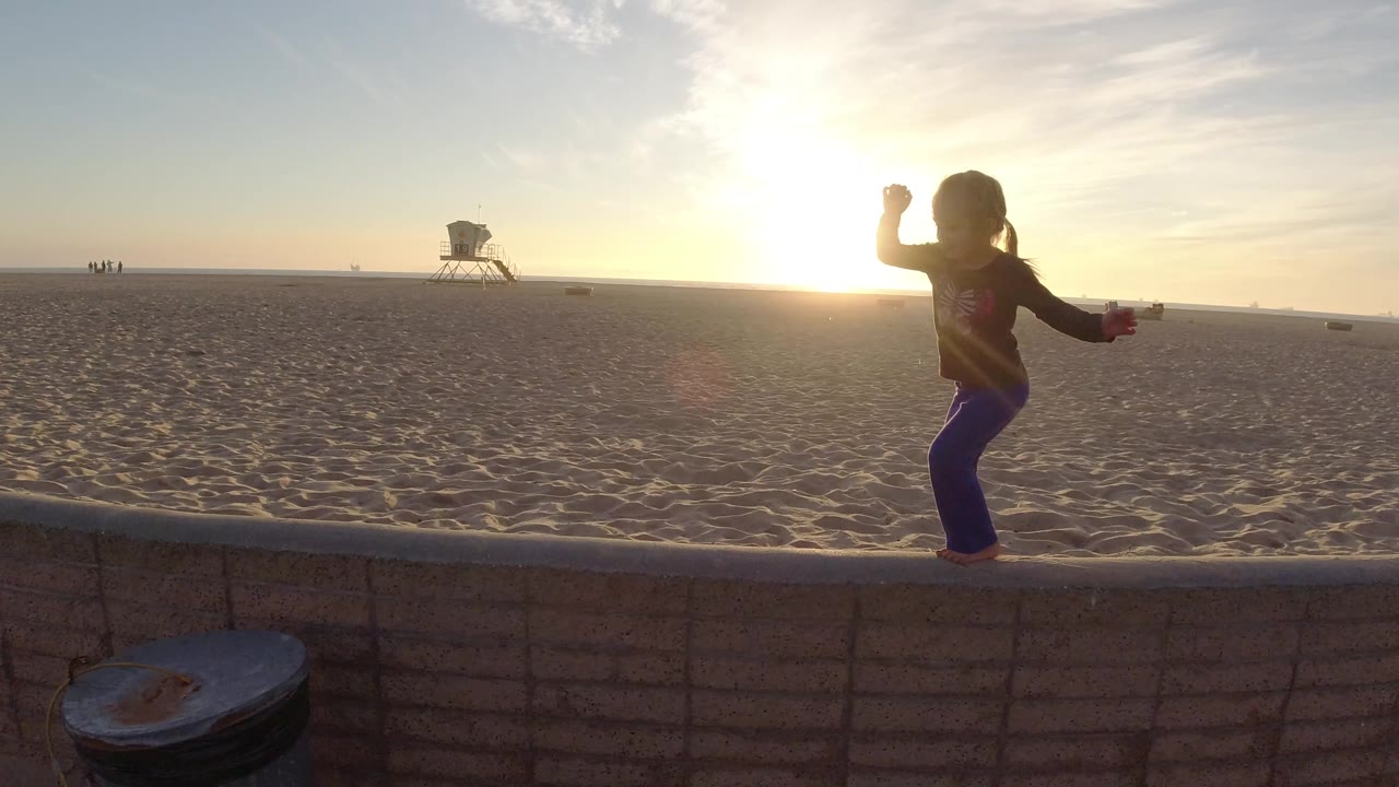 A young girl stands on a low wall at Bolsa Chica beach with the low sun behind her, open sand ahead, and a distant lifeguard tower near the horizon.