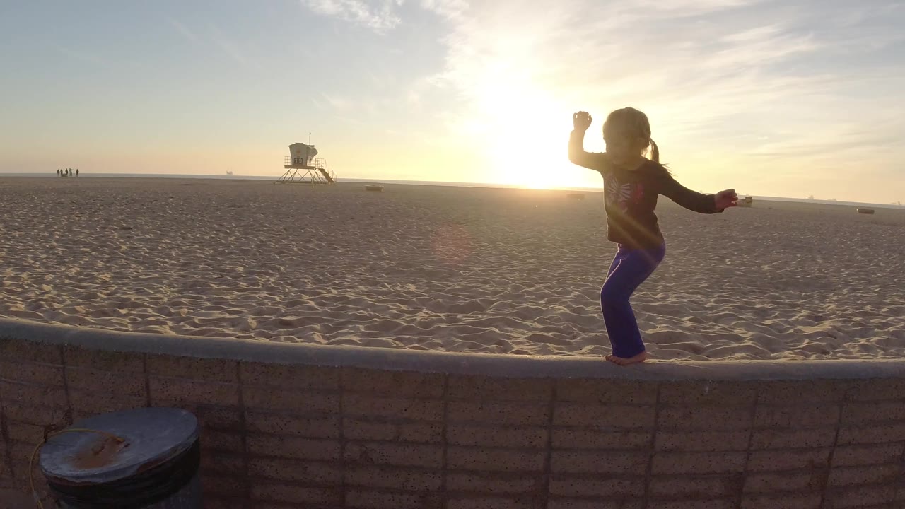 The child shifts along the low wall at Bolsa Chica with one arm raised, while sunset glare washes the sand and the lifeguard tower remains in the distance.