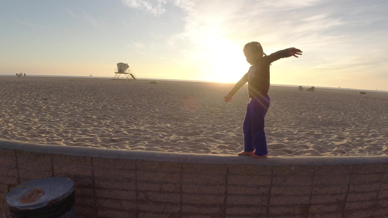 The girl appears near the right side of the Bolsa Chica beach frame with arms extended, backlit by the low sun over a broad sandy background.