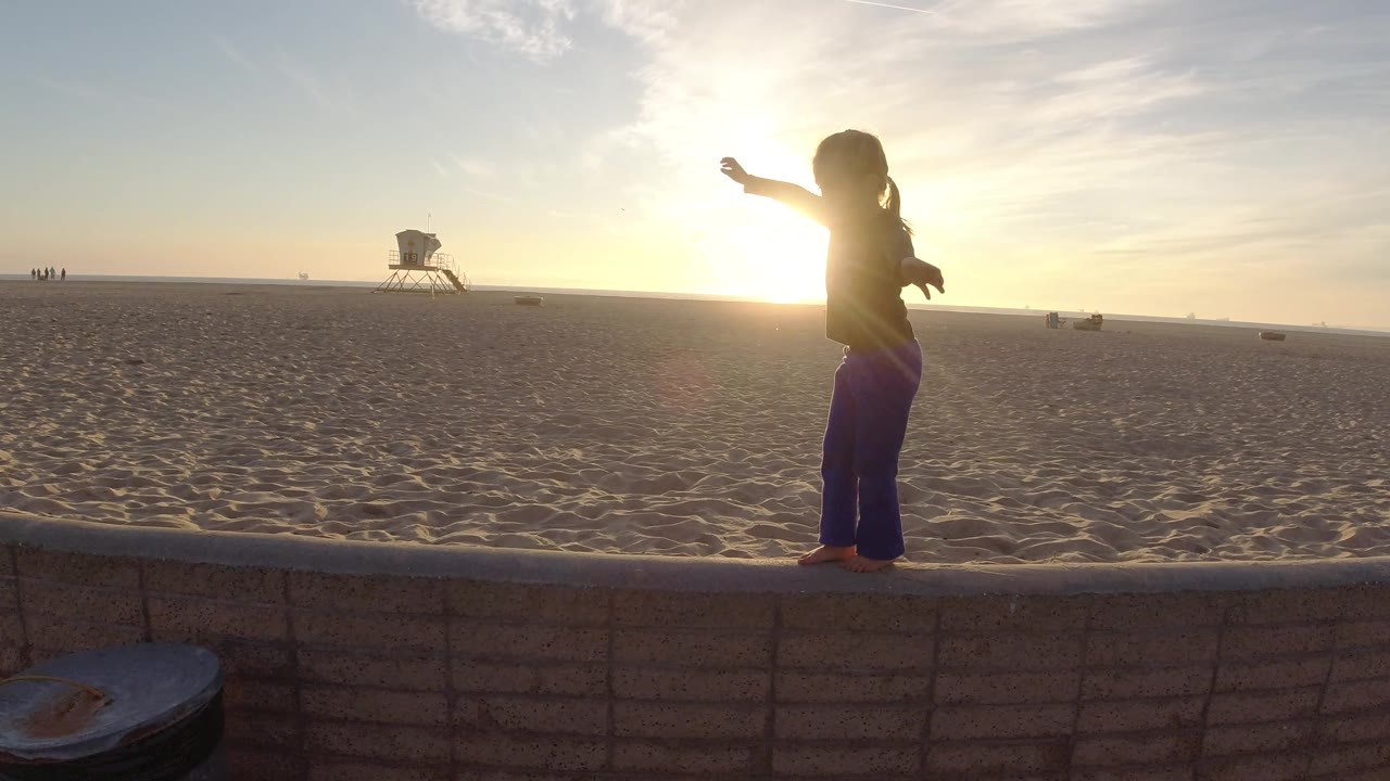 Backlit girl on beach wall with lifeguard tower in the distance.