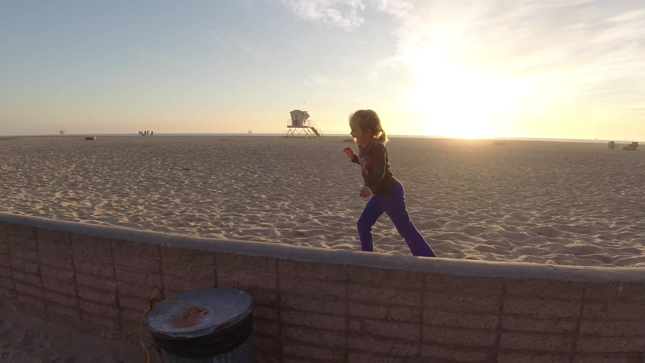 Young girl runs to the right along the low beach wall at Bolsa Chica, backlit by the low sun with open sand and a lifeguard tower beyond.