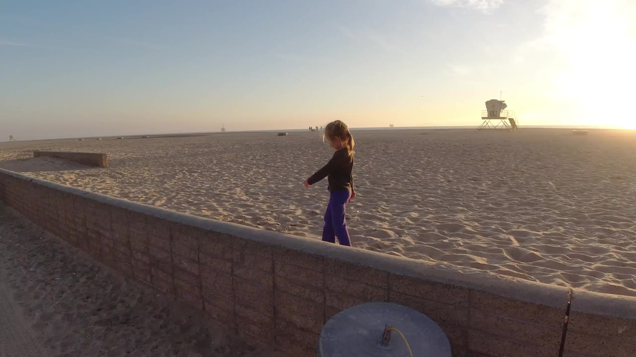 At Bolsa Chica, California, a young girl walks near a low wall with wide rippled sand and a distant lifeguard tower under bright low sun.