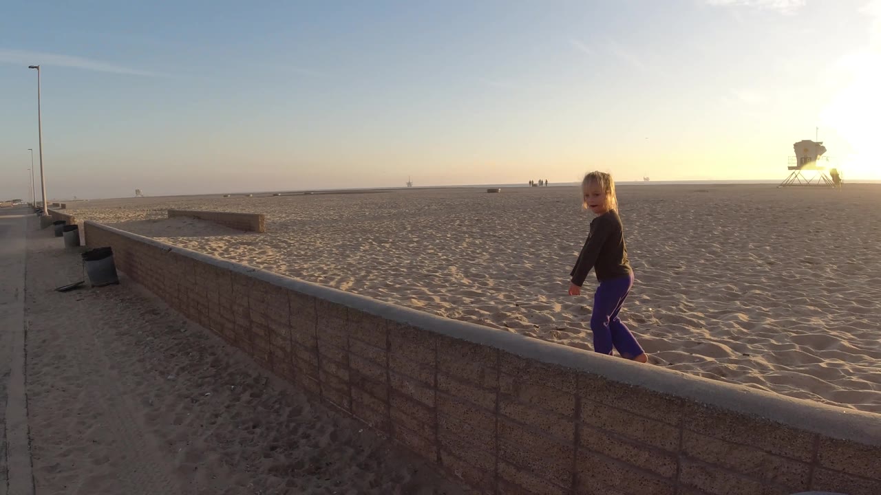 At Bolsa Chica, California, the child appears near the right edge while the low wall, lamps, sand, and distant lifeguard tower stretch across the bright beach view.