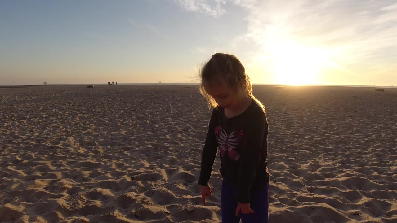 A child in dark clothing stands near the foreground on Bolsa Chica beach sand, backlit by the low sun with a wide empty horizon behind them.