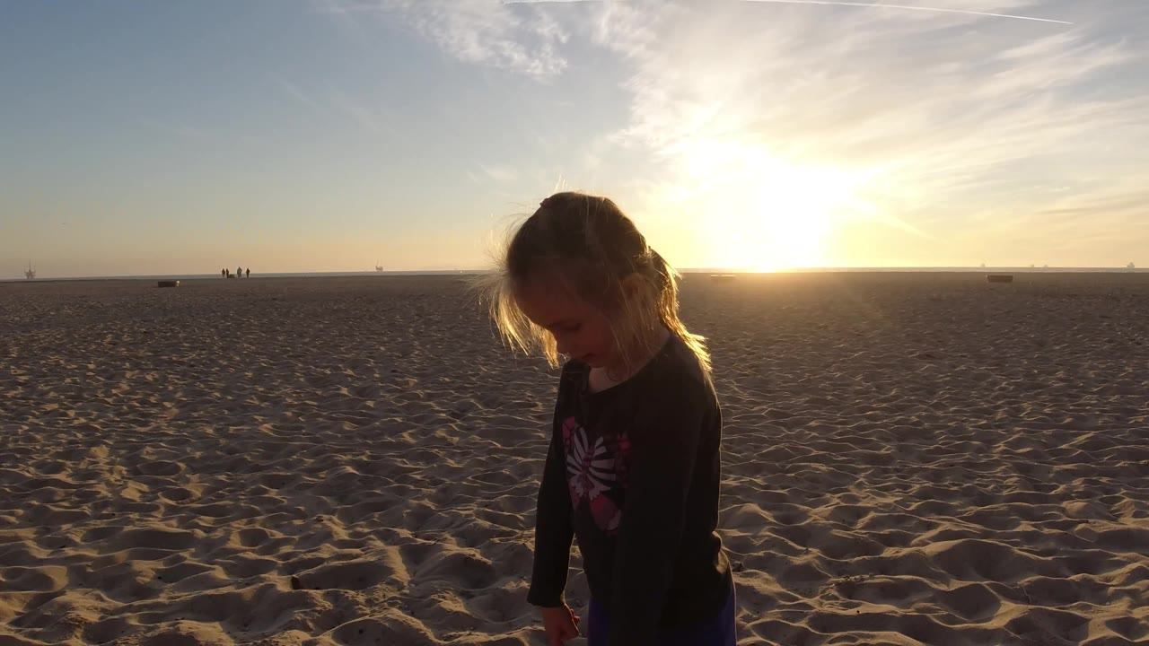 A child appears near the right side of the Bolsa Chica beach frame, with broad sand in front, distant figures on the horizon, and the sun glowing behind.
