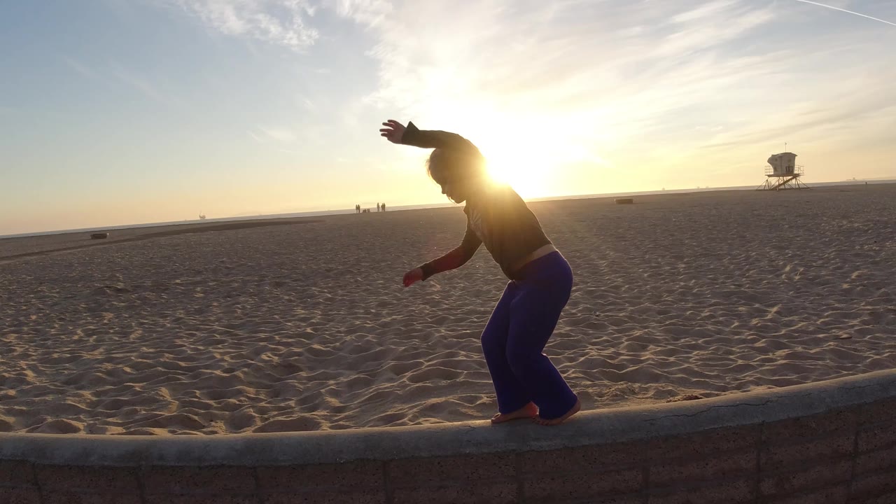 At Bolsa Chica, California, the toddler keeps arms raised for balance on the low wall as the sun sits directly behind the child and the sandy beach stretches into the background.