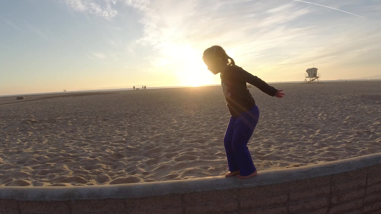 At Bolsa Chica, California, the toddler stands more upright on the low wall, with the beach, low sun, distant figures, and a lifeguard tower visible across the sand.