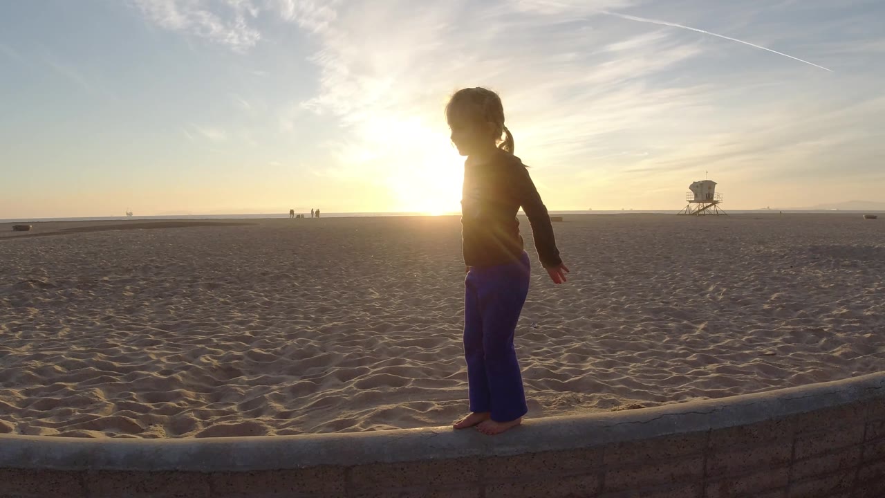 Backlit toddler steps along the edge of a low wall near sand.