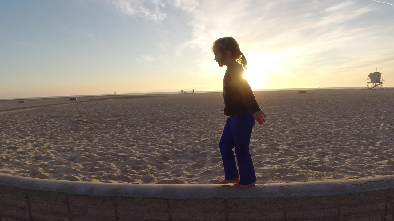Young girl in blue pants walks barefoot along a low wall at Bolsa Chica Beach, with sun glare behind her and a lifeguard tower in the distance.