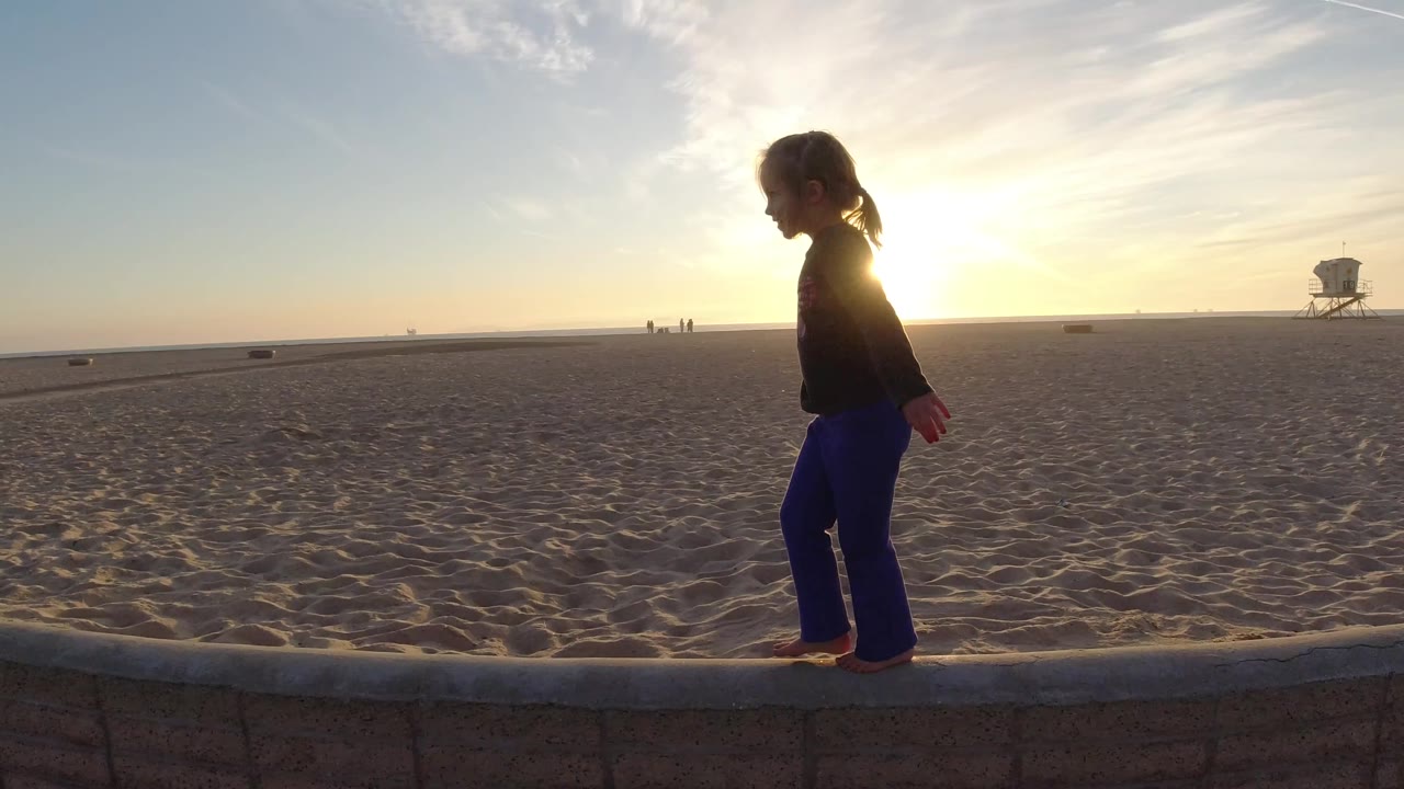 At Bolsa Chica Beach, the girl continues along the low concrete wall, silhouetted by the sun with open sand and a distant tower behind her.
