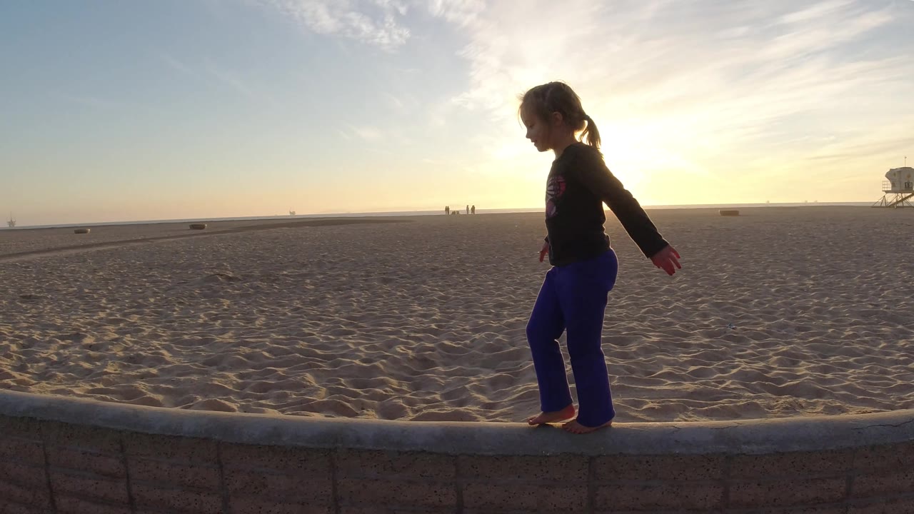 The child is farther right on the low wall at Bolsa Chica Beach, with her arms slightly out and the lifeguard tower set near the right edge of the horizon.