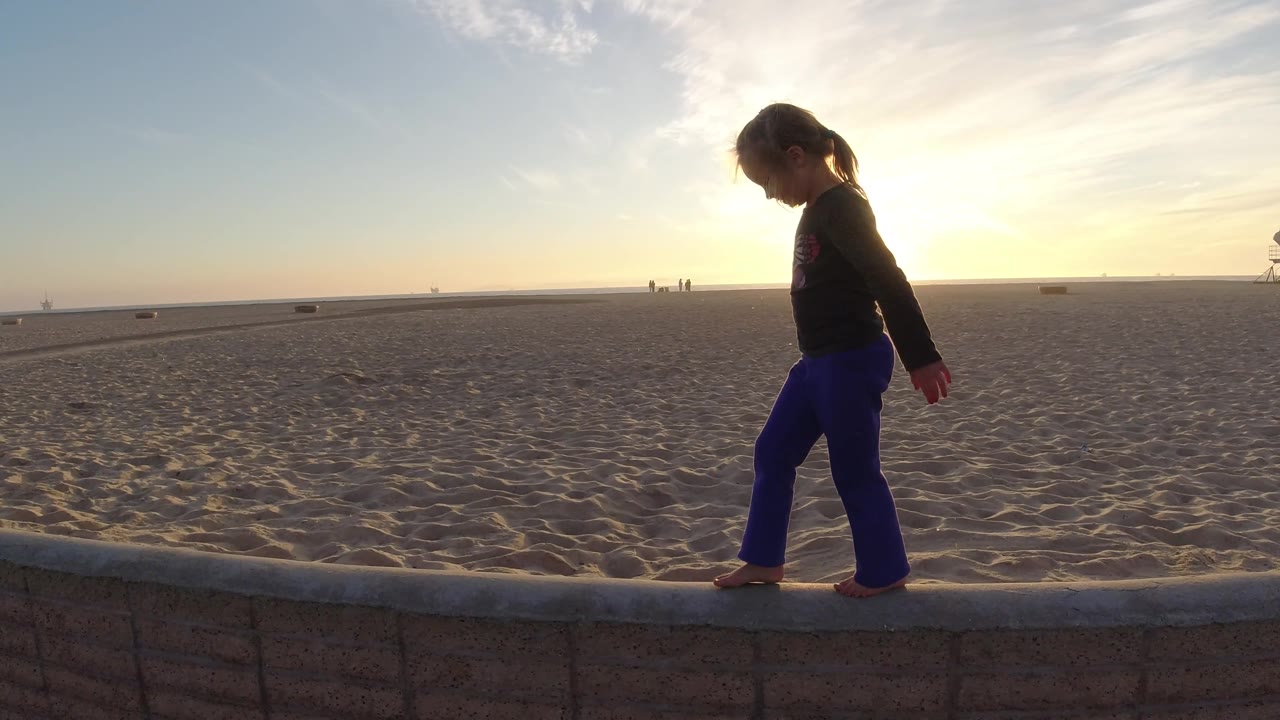 Girl walks barefoot along a beach wall with sunset backlight.
