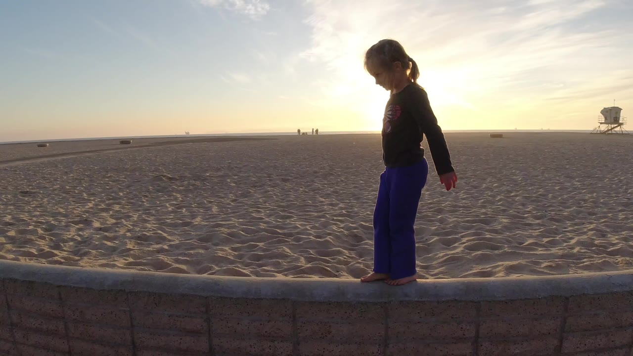 A young girl stands in profile on a low concrete beach wall in Bolsa Chica, California, with open sand, a lifeguard tower, distant figures, and bright sun behind her.