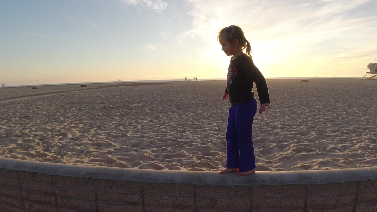 The girl appears farther right on the low wall in Bolsa Chica, California, with her side profile outlined against the bright sky and the lifeguard tower near the far right edge.