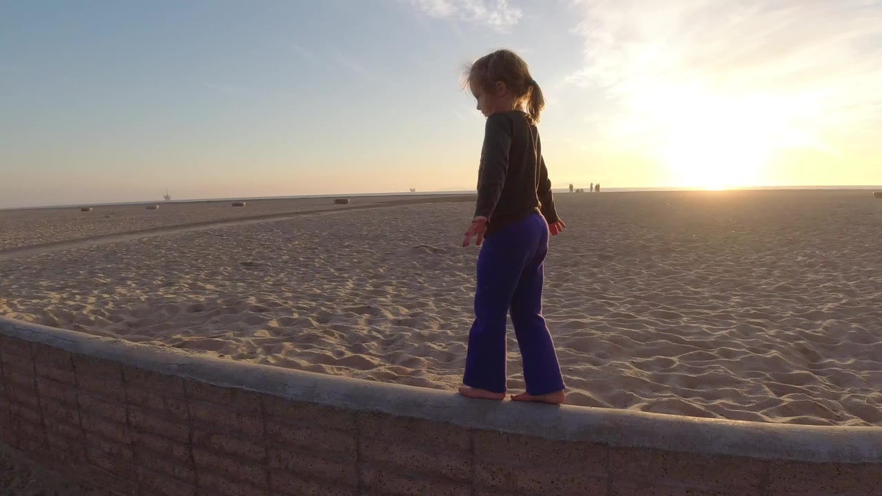 A young girl stands barefoot on a low wall at Bolsa Chica, California, with open sand, a flat ocean horizon, and the low sun behind her.