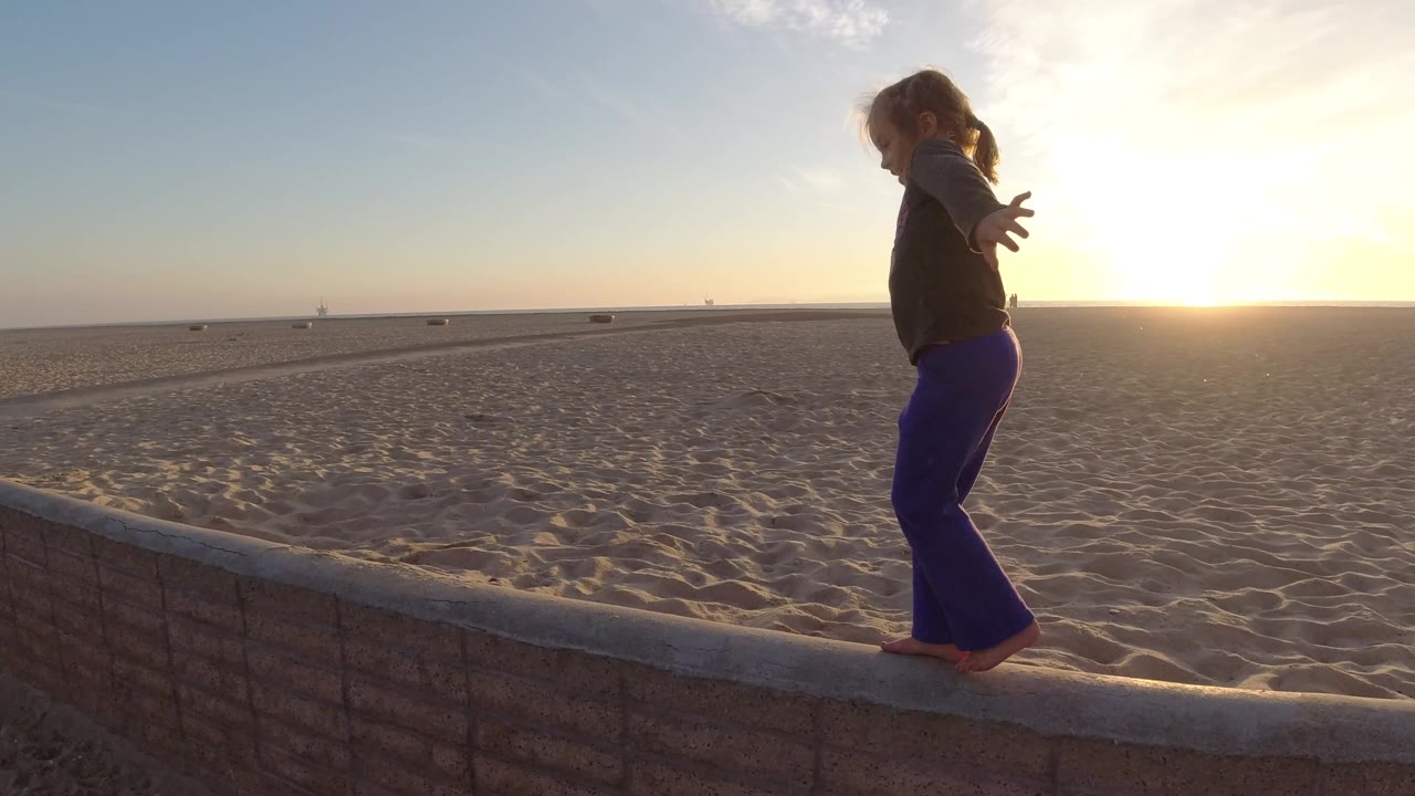 The child balances on the wall with one arm extended, silhouetted by the low sun over the sand and horizon at Bolsa Chica, California.
