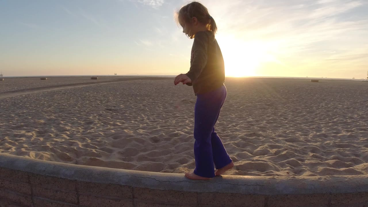 At Bolsa Chica, California, a young girl walks barefoot along a low concrete edge with open sand and a bright low sun behind her.