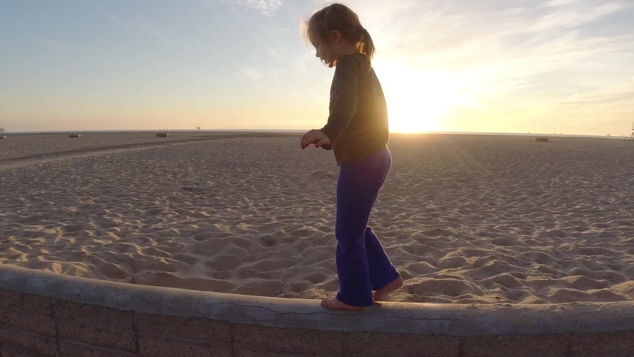 The girl continues along the concrete edge at Bolsa Chica, California, with the sun flaring near her torso and a wide sandy beach behind her.