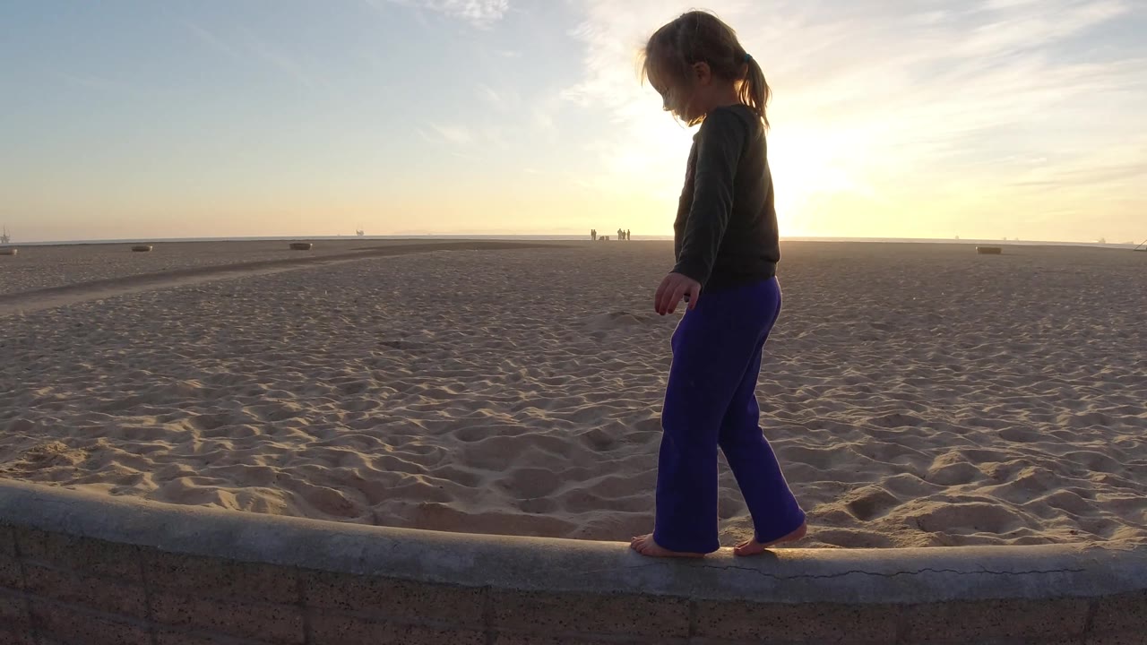 At Bolsa Chica, California, the child is farther along the ledge, silhouetted against open beach, pale sky, and the low sun near the horizon.