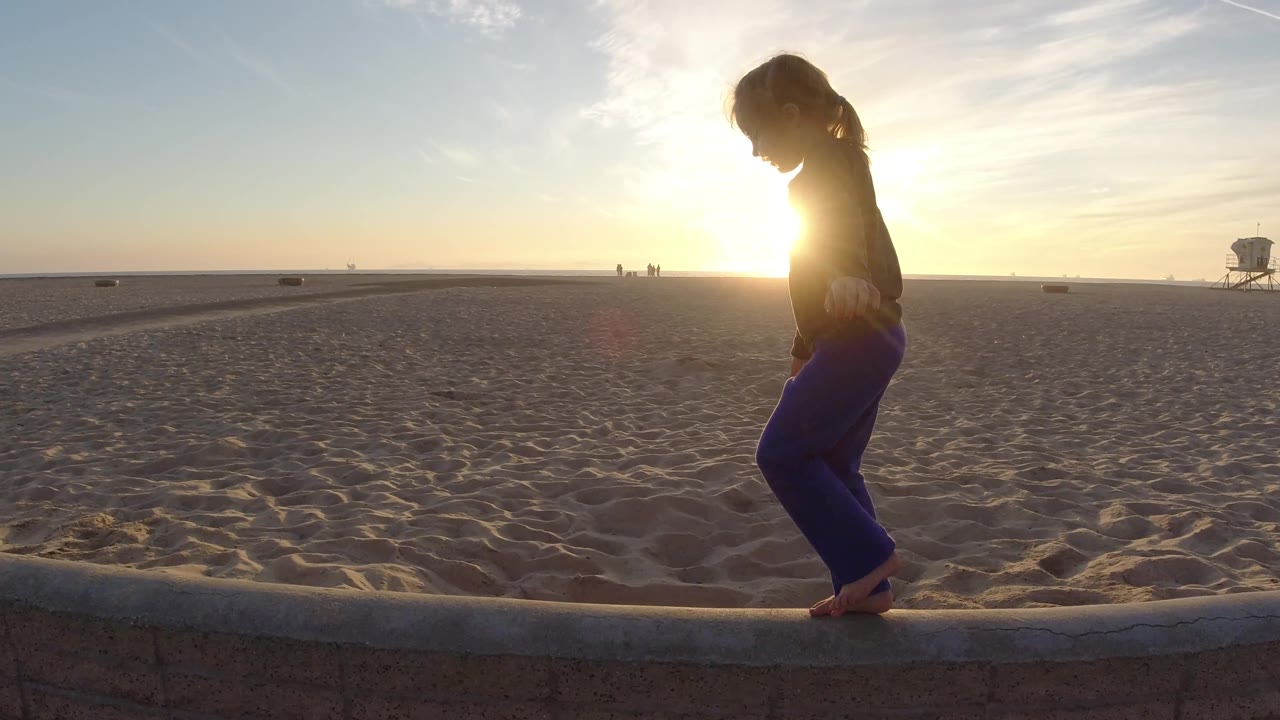 Backlit girl balancing on a beach ledge near sunset.