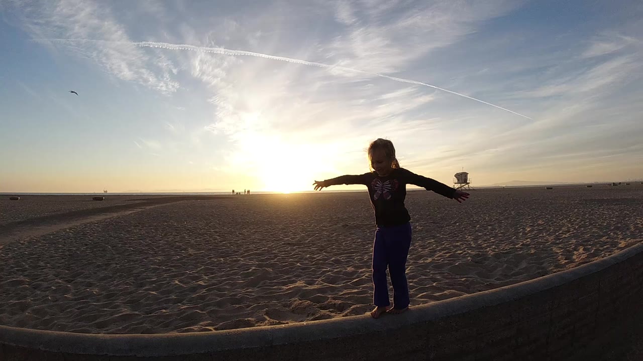 The child remains on the curved beach edge at Bolsa Chica, California, with one arm reaching toward the sunset and the lifeguard tower visible to the right.