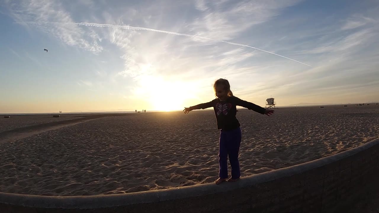 A child stands slightly right of center on the beach edge in Bolsa Chica, California, silhouetted by the low sun with open sand and a lifeguard tower behind.