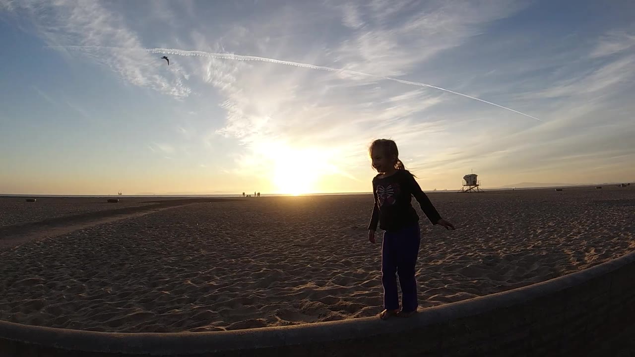 A small girl stands farther right in the wide Bolsa Chica beach view, with the sun low at left, textured sand across the frame, and thin cloud lines above.