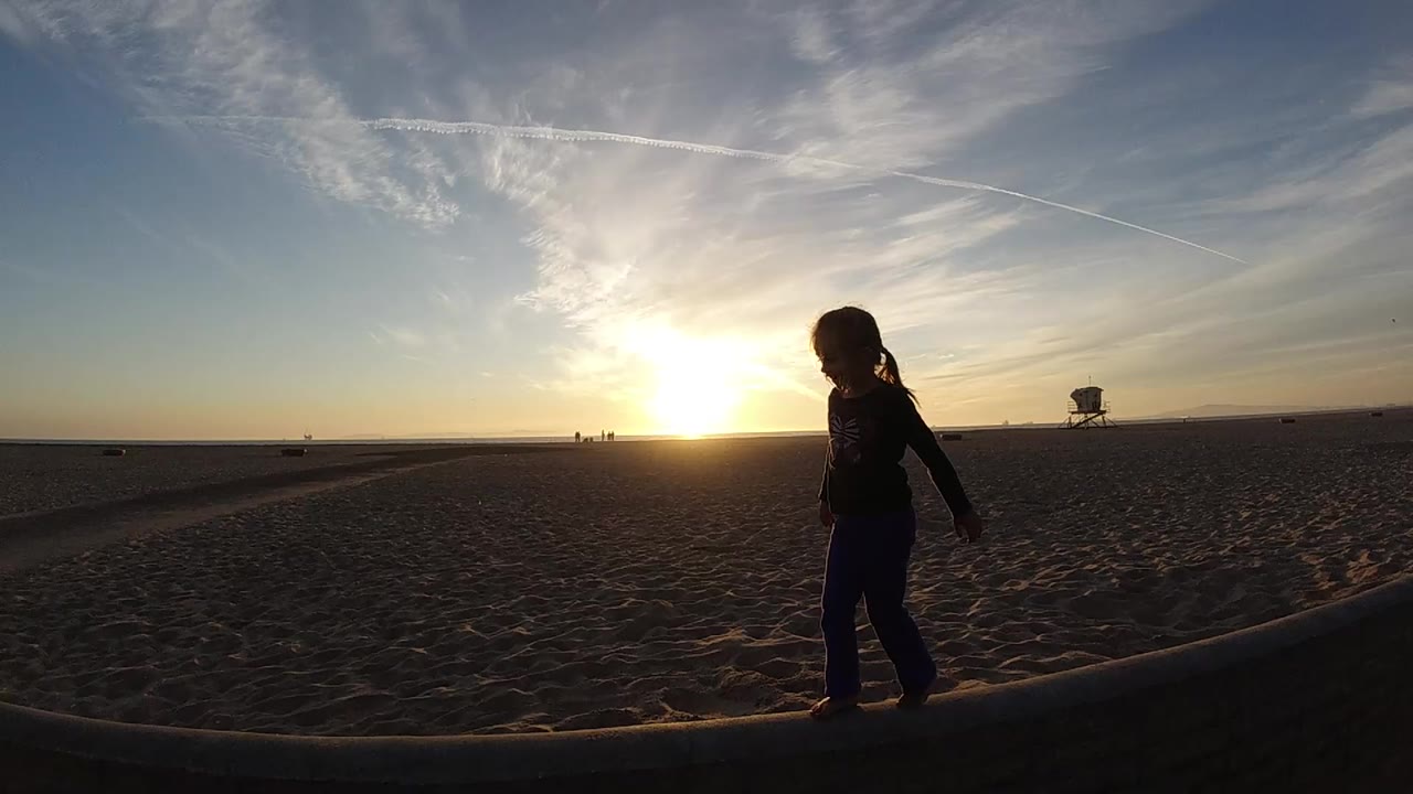 At 0:00 in Bolsa Chica, California, a young girl appears in dark silhouette on a wooden beam as the sun sits low over the sandy horizon.