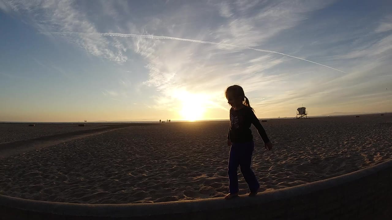 At 0:01 in Bolsa Chica, California, the silhouetted girl is centered on the beam while bright sun glare and diagonal jet trails fill the sky behind her.