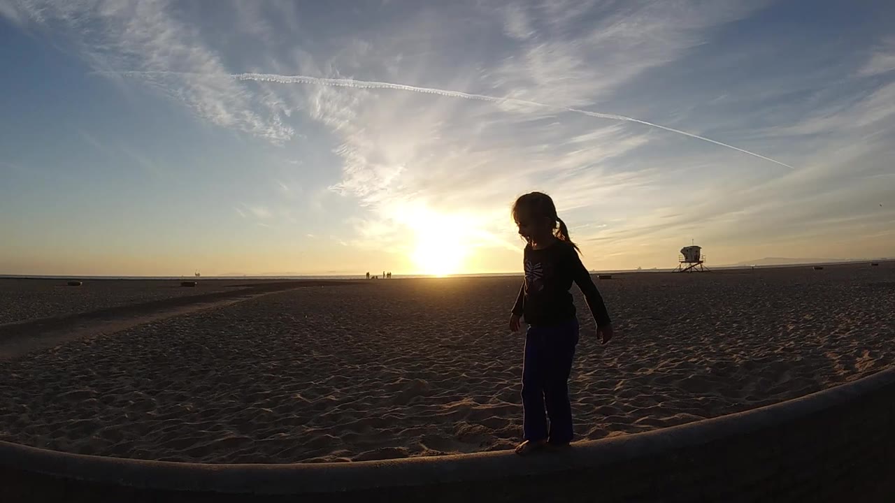 At 0:02 in Bolsa Chica, California, the girl has moved toward the right side of the beam, leaving more open sand and sunset sky across the left of the frame.
