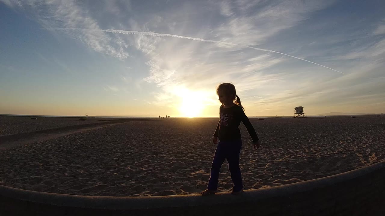 A silhouetted child walks near the foreground edge of a Bolsa Chica beach at sunset, with the sun low to the left and a lifeguard tower far in the background.