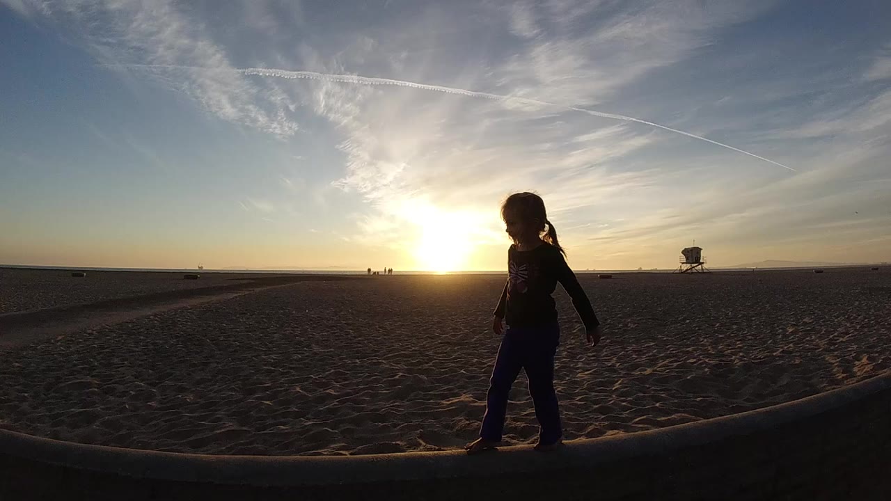 At 0:01, the child silhouette is closer to center on the Bolsa Chica beach, with long dark sand texture in front and thin clouds above the low sun.