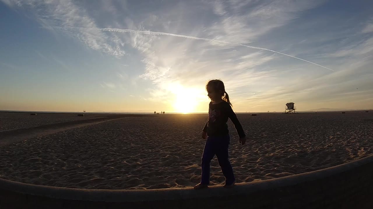 At 0:02, the silhouetted child has moved farther right across the foreground at Bolsa Chica, while the lifeguard tower sits small on the distant beach.