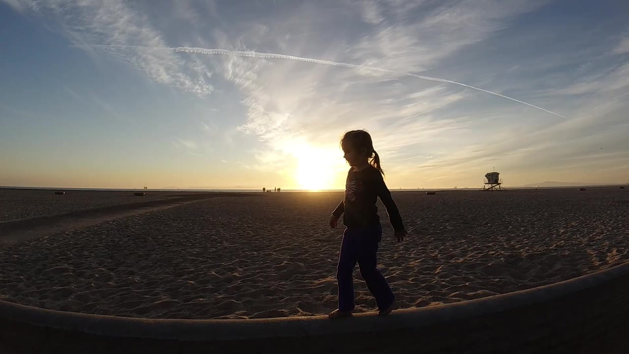 Backlit child standing or walking on beach edge with sun near the horizon.